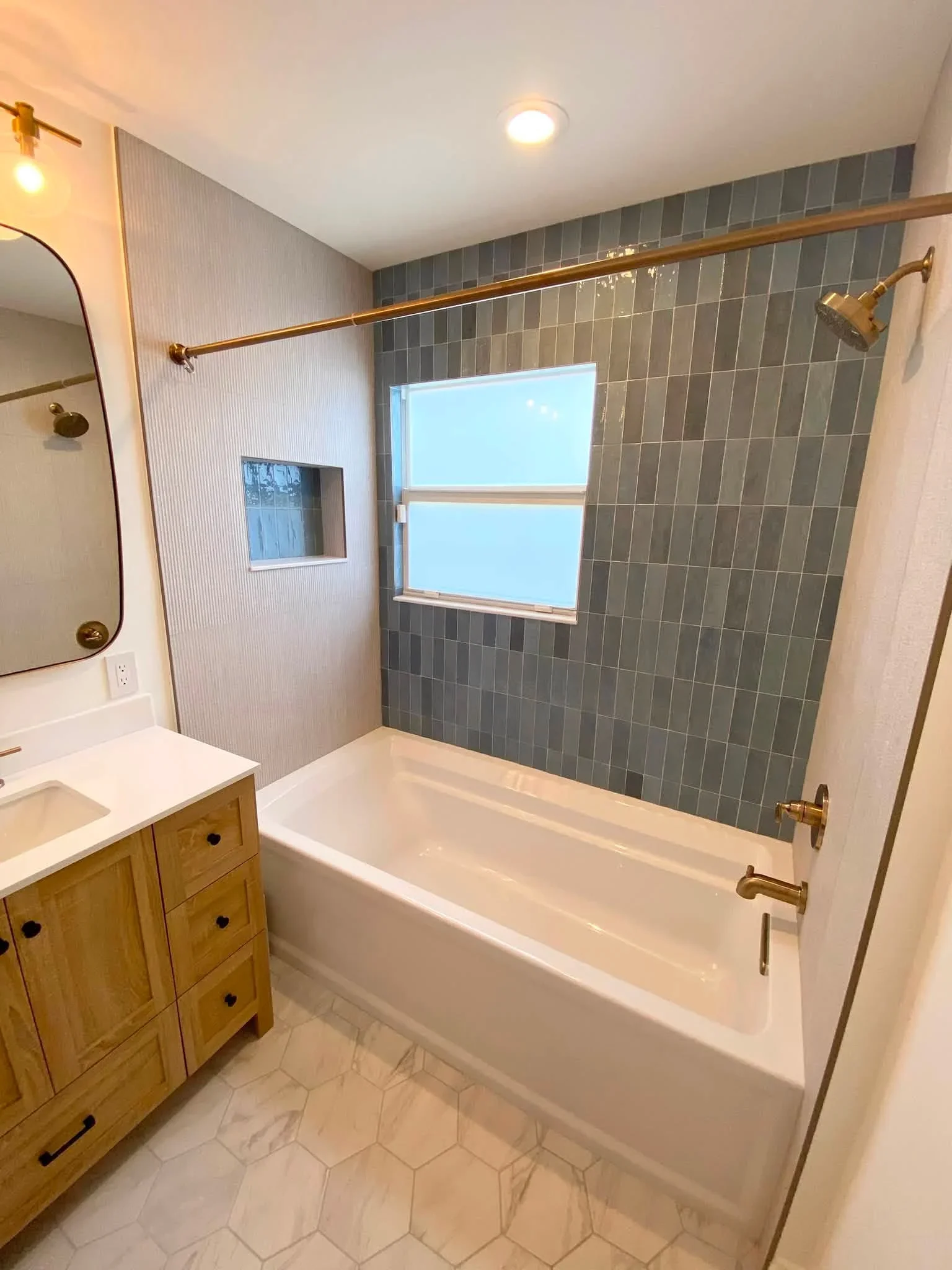Bathroom with white tub, charcoal tile surround, glass shower enclosure, wood vanity, and brass fixtures